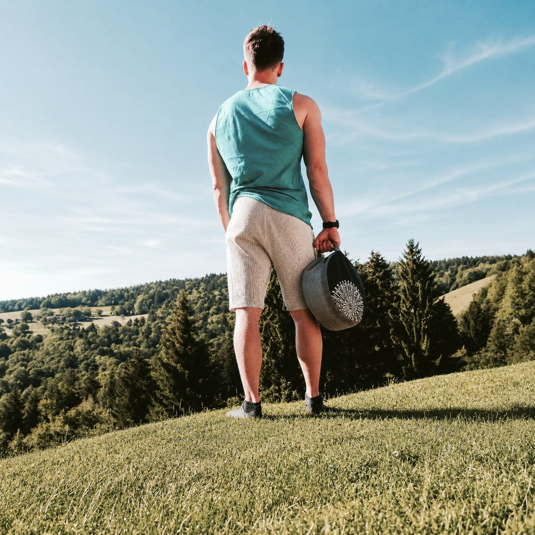 Man holding a grey AKASHA meditation cushion by its built-in handle, standing on a grassy hill overlooking a lush forest landscape.