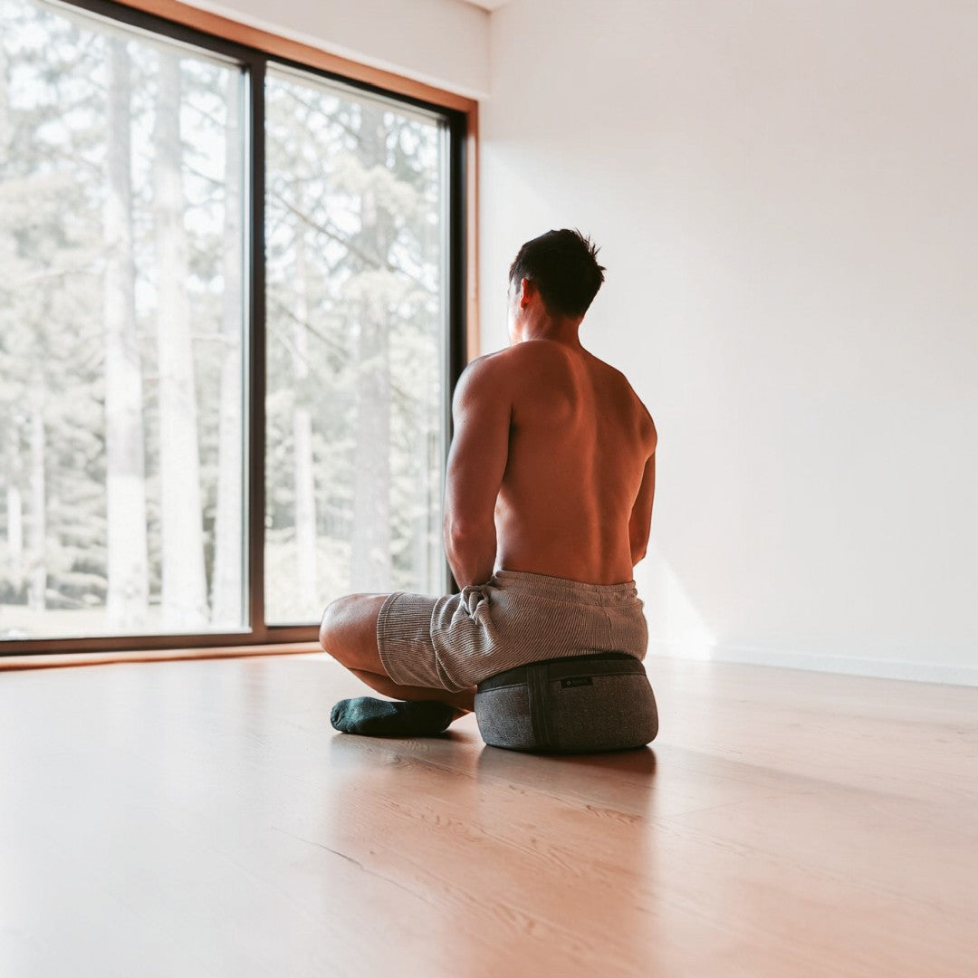 Side view of a man sitting in a cross-legged position on a grey AKASHA meditation cushion, practicing mindfulness in a bright room with a large window.