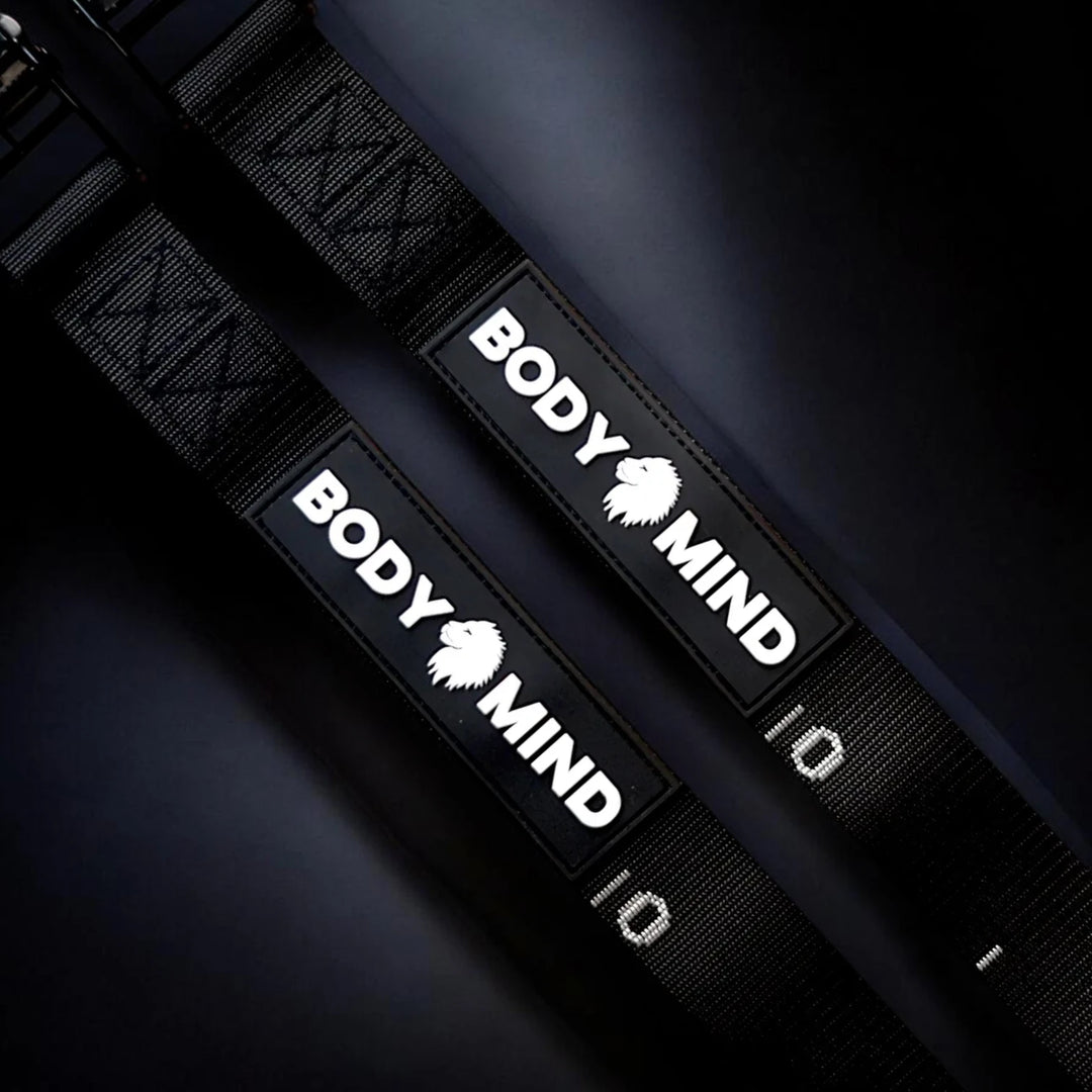 Two black gymnastic rings adjustable straps with white 'BODY MIND' branding, shown horizontally against a dark black background.